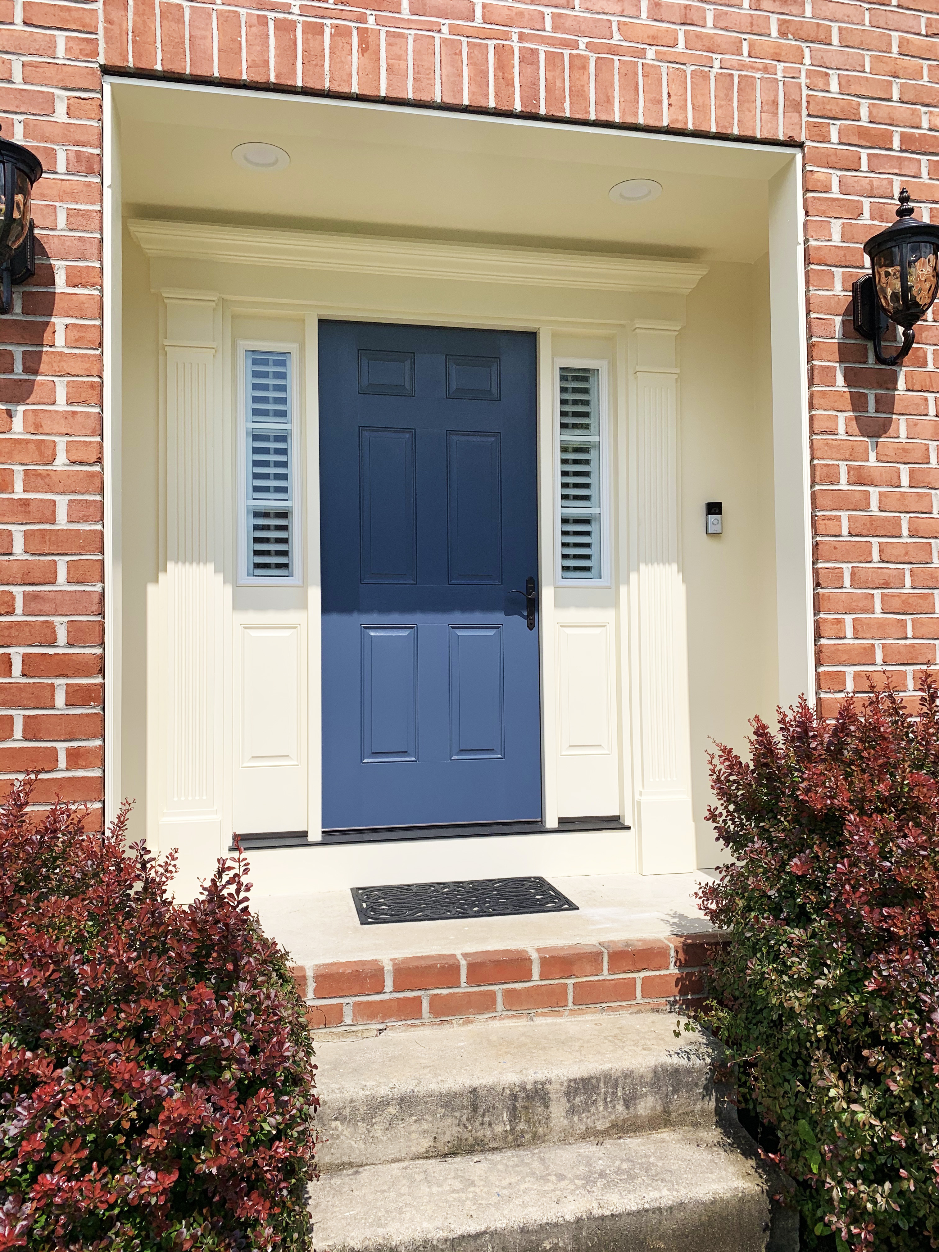 Residential entryway remodel featuring a navy blue front door with cream colonial trim, brick exterior, decorative lighting fixtures, and manicured landscaping for classic curb appeal.